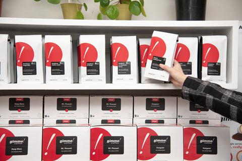 A person's arm reaching up and grabbing a bag of Gimme coffee beans off of a shelf full of bags of coffee and boxes of k-cups.